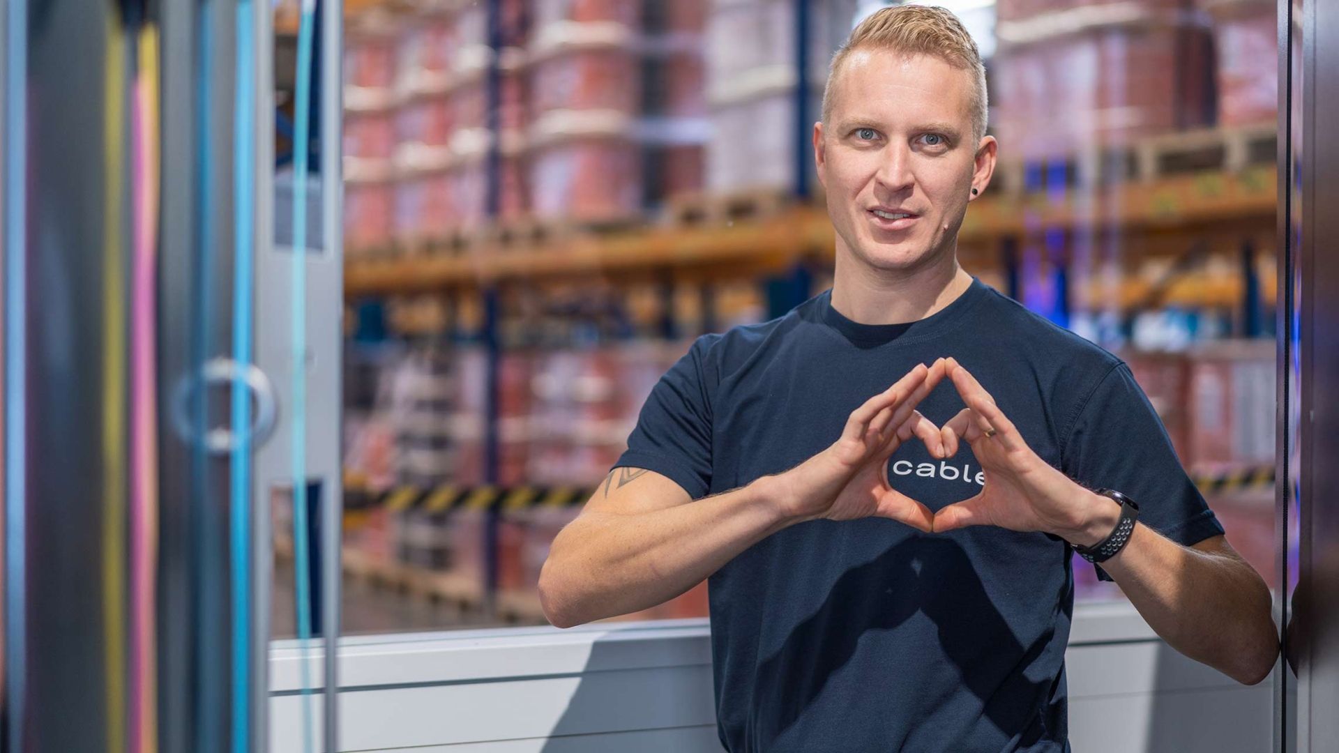 cablex Mitarbeitender formt mit der Hand ein Herz über den cablex-Schriftzug auf seinem T-Shirt.